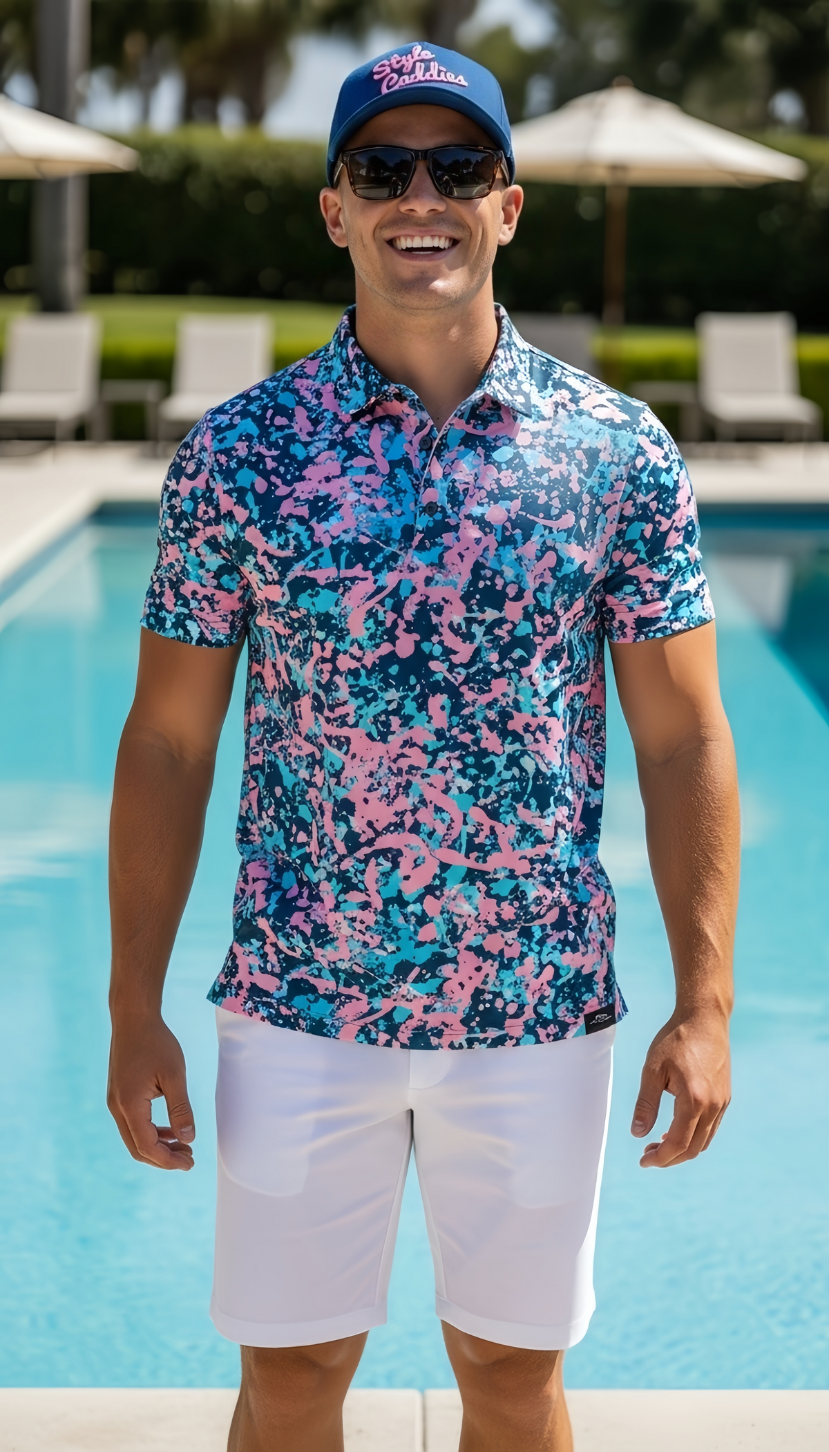 Smiling model wearing the Style Caddies Pinkulator splatter print golf polo with white shorts and a matching Purpulator trucker hat, poolside